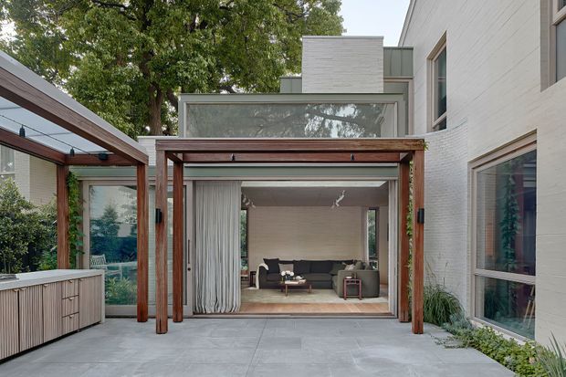 Contemporary courtyard viewed from an outdoor entertaining area, featuring a timber-framed pergola with climbing greenery, pale concrete paving, a built-in outdoor kitchen or storage unit at left, and a glazed living room opening directly onto the patio. The residence has white painted brick walls, large windows, and a projecting upper-level glazed clerestory that brings light into the interior.