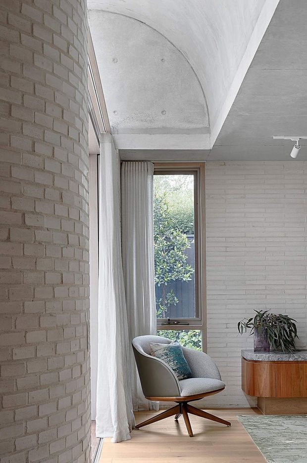 A bright, narrow interior sitting area with exposed white-painted brick walls, a curved concrete ceiling feature, timber flooring, and a tall window overlooking greenery. A mid-century style upholstered lounge chair sits beside a curved wooden sideboard with stone or terrazzo-like top, with soft white curtains and a small rug completing the space.