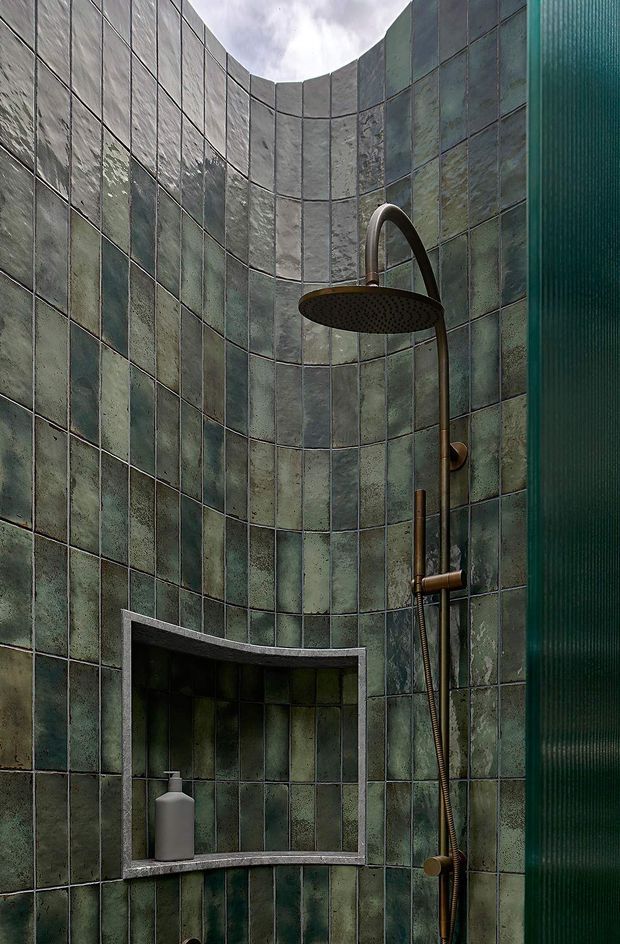 Close-up view of a curved shower enclosure lined with vertically oriented, mottled green and grey ceramic tiles. A large round rainfall showerhead and handheld shower are mounted on a dark bronze or brass system, with a recessed tiled niche containing a bottle. A translucent ribbed green glass screen is visible at the right edge.