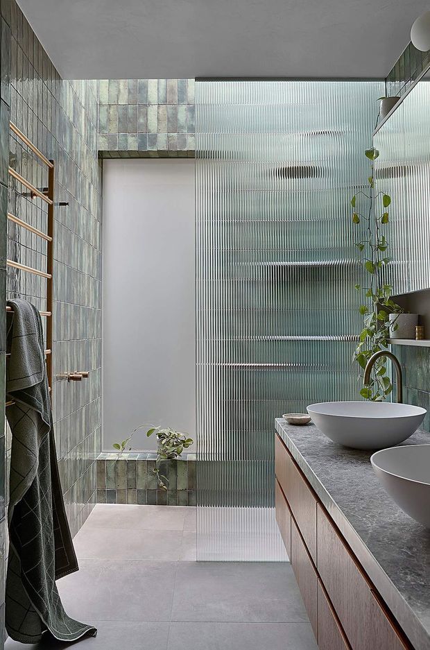 A contemporary bathroom with a walk-in shower, twin vessel basins on a timber vanity, grey stone countertop, and muted green-grey wall tiles. A ribbed translucent glass screen provides privacy and texture, while a timber heated towel rail and hanging plants add warmth and natural detail. The space is softly lit with a calm, refined palette.