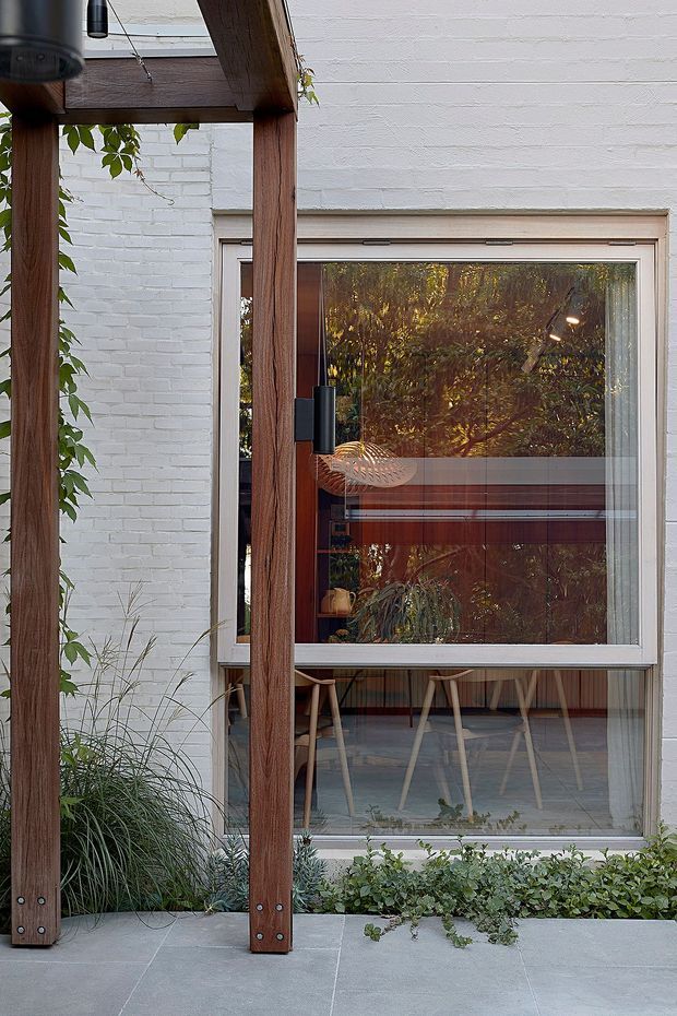 A close exterior view of a contemporary residential facade, featuring white-painted brick walls, a large glazed window or door opening, and a timber pergola with substantial vertical posts. The glazing reflects surrounding foliage while revealing a timber-lined interior with stools and a dining or work counter. Grey tiled paving, climbing greenery, and low planting frame the facade.