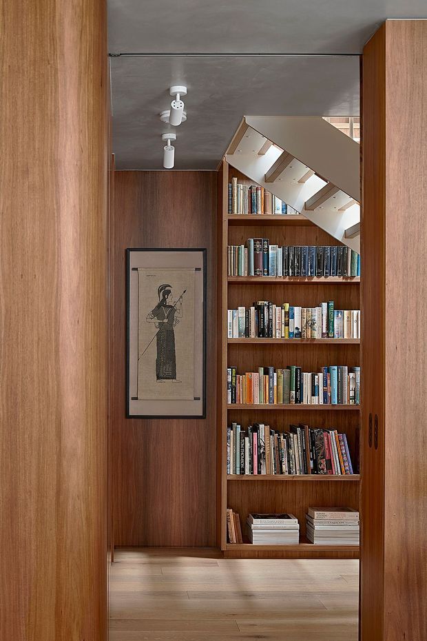 A narrow, highly crafted timber-lined interior passage or study nook, viewed through open timber doors. Floor-to-ceiling built-in timber shelving is filled with books, with a framed artwork on the timber wall and a compact white track-light system on the dark ceiling. A white stair structure crosses above the shelving, suggesting a space beneath or beside a staircase. The overall composition is warm, refined, and strongly joinery-focused.