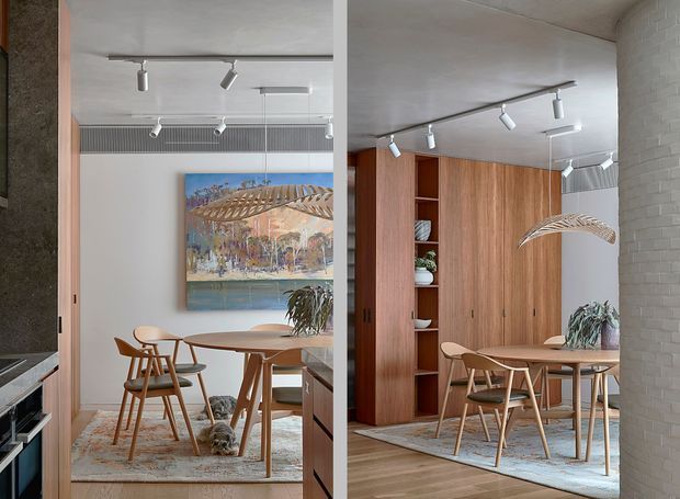 A refined dining area viewed through a partially open doorway, featuring a round timber dining table with matching mid-century-style wooden chairs, a patterned area rug, warm timber built-in cabinetry with open shelving, track lighting, a large framed artwork, and a white painted brick wall. The image has a split or mirrored editorial composition showing the room from two closely related views.