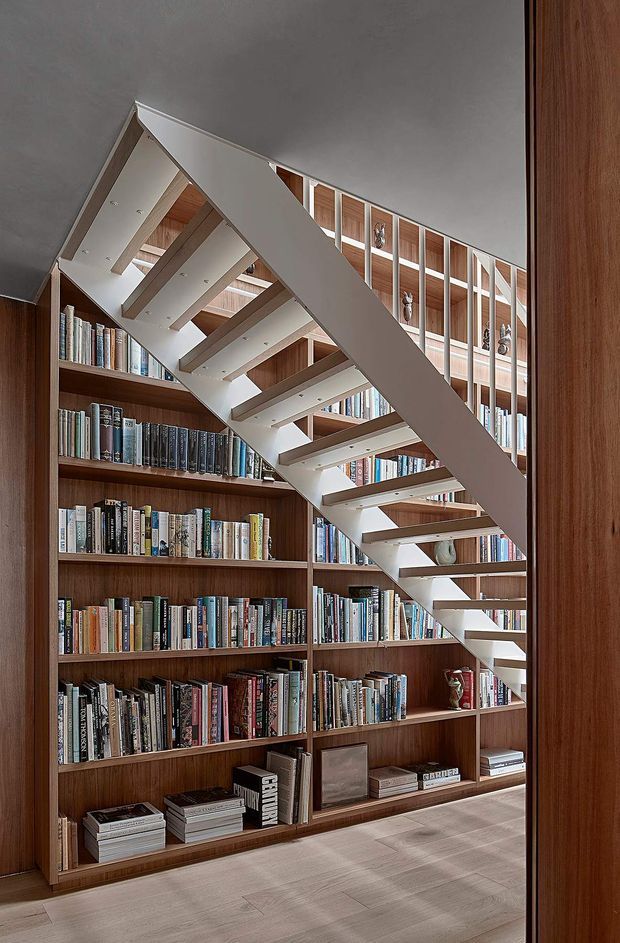 A refined library space viewed through a timber-framed opening, featuring extensive floor-to-ceiling dark timber bookcases filled with books and objects. A sculptural open-riser staircase with white stringers and treads crosses diagonally through the room, creating a strong architectural focal point. The room has a warm timber-and-white palette, a smooth light-toned floor, and a tall, airy vertical composition.