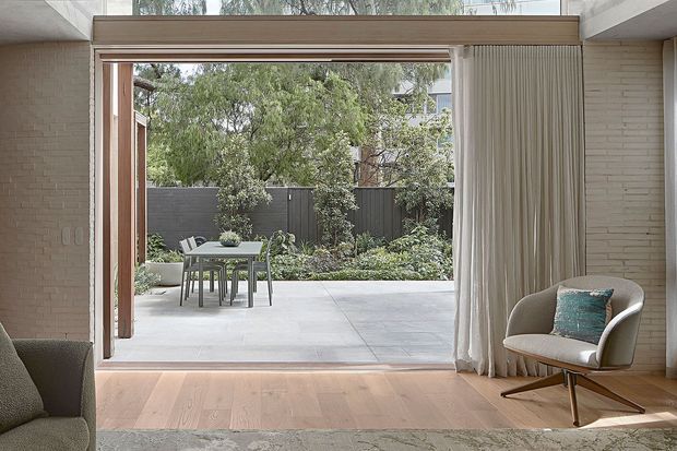 A calm, light-filled interior looking through wide open timber-framed glazing and folding doors onto a paved landscaped courtyard. The room has pale timber flooring, light textured brick walls, full-height curtains, a patterned rug, and a mid-century-style lounge chair. An outdoor dining setting and planted garden are visible beyond.