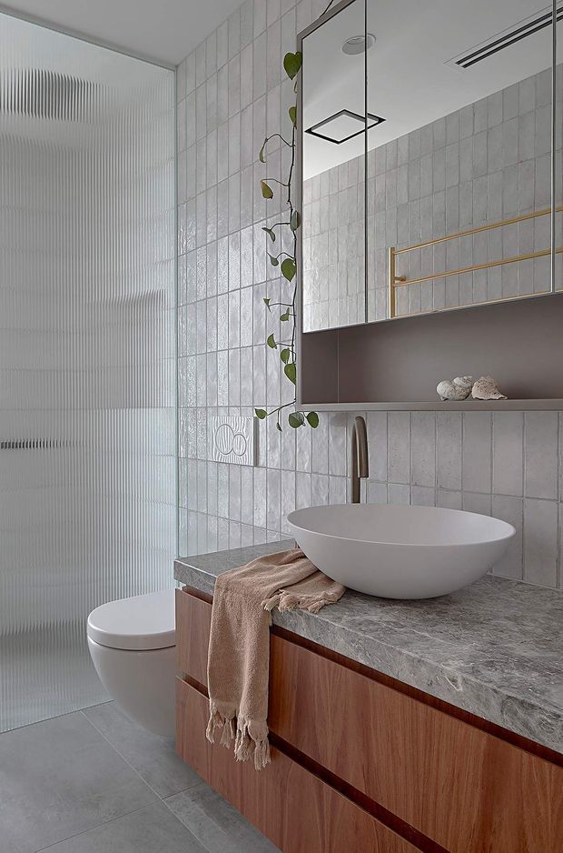 A refined contemporary bathroom featuring a floating timber vanity with integrated drawers, a grey stone benchtop, a white vessel basin and brushed metal tapware. Pale vertically textured wall tiles and a ribbed translucent glass shower screen create a softly tactile backdrop. A large mirrored cabinet, warm brass towel rail, trailing plant and neutral linen add understated warmth.