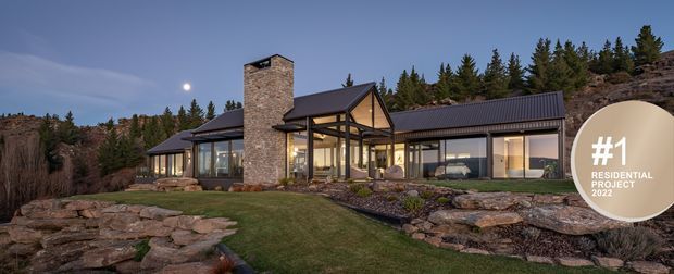 Exterior dusk view of a large contemporary mountain residence set into a rocky, landscaped site. The house has extensive floor-to-ceiling glazing, dark metal pitched roof forms, warm timber framing, and a prominent tall stone chimney or masonry feature. Coniferous trees and exposed rock surround the property, while a circular award graphic overlays the right side of the image.