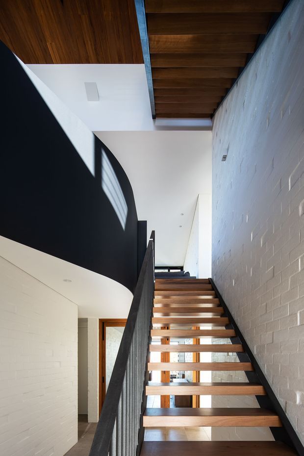 Interior view of a modern stairway with open-riser timber stairs, black metal balustrades, white-painted brick walls, and a warm timber-lined ceiling. Strong natural light enters through glazing near the stair landing, creating high contrast and highlighting the clean, angular architectural composition.
