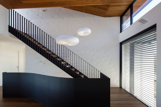 A bright contemporary interior featuring a dramatic black staircase with slender vertical metal balustrades, white painted brick walls, dark timber flooring, and a warm timber-lined ceiling. Large glazed openings with horizontal blinds bring in abundant daylight, while sculptural white pendant lights hang over the stair and landing area.