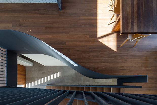 A top-down architectural view of a contemporary interior stairway. Dark, sculptural stair treads and a sweeping curved balustrade frame a warm dark timber floor, with a concrete-toned wall and strong sunlight creating linear shadows. A dining or work area with timber joinery and yellow chairs is visible at the upper right.