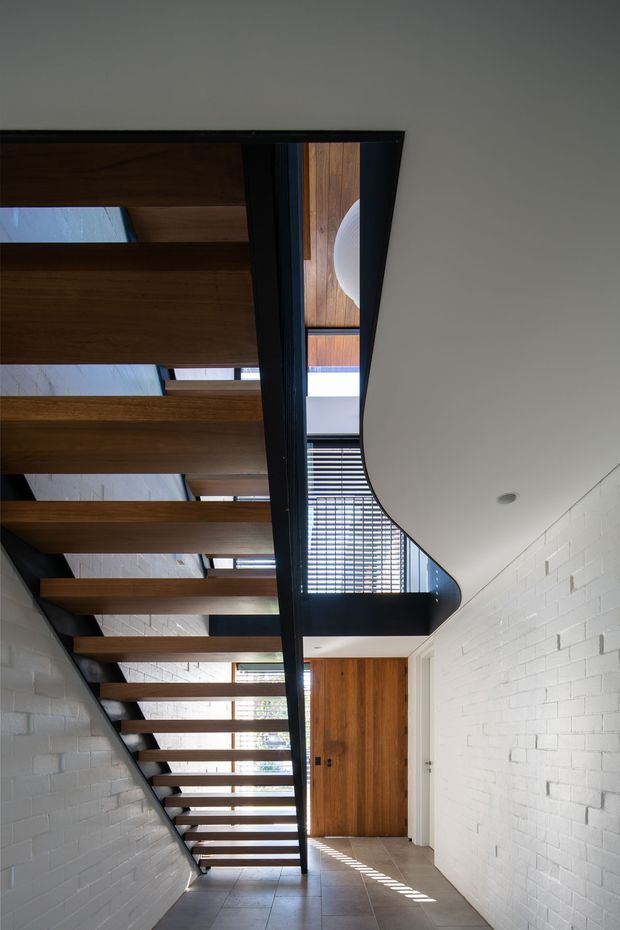 A contemporary interior stairway viewed from below, featuring an open-riser timber staircase with dark metal stringers and balustrade. White-painted brick walls, exposed timber ceiling beams, roof glazing or skylights, and a curved white ceiling create a dramatic, light-filled circulation space. A timber door and tiled floor are visible beneath the stair.