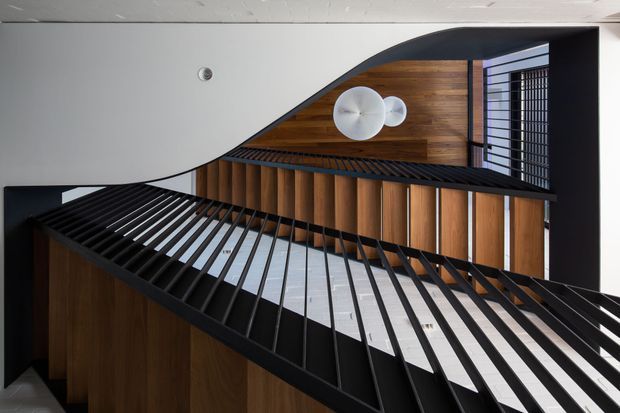 Interior architectural view of a dramatic curved stairway or landing with black metal balustrades, warm vertical timber wall panelling, and a sculptural white curved ceiling. A large round pendant light hangs above, with strong geometric lines and contrasting light and dark finishes.