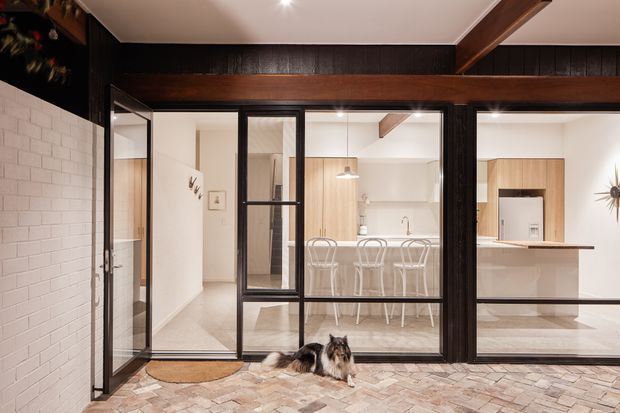 Interior view from a covered outdoor or entry area through black-framed glazed doors into a bright, modern kitchen and dining space. The kitchen features light timber cabinetry, a central island with bar seating, pale benchtops, white walls, and exposed dark timber beams. A white painted brick wall and patterned tiled floor are visible in the foreground, with a dog near the threshold.