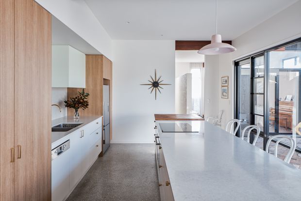 Bright, open-plan kitchen with extensive light timber cabinetry, a long pale stone island with integrated seating, and a compact galley-style work area. The space includes a stainless-steel sink, white lower cabinets, polished concrete flooring, a sculptural pendant light, and large black-framed glazed doors opening to an outdoor area. A mid-century starburst wall clock and restrained furnishings add character.