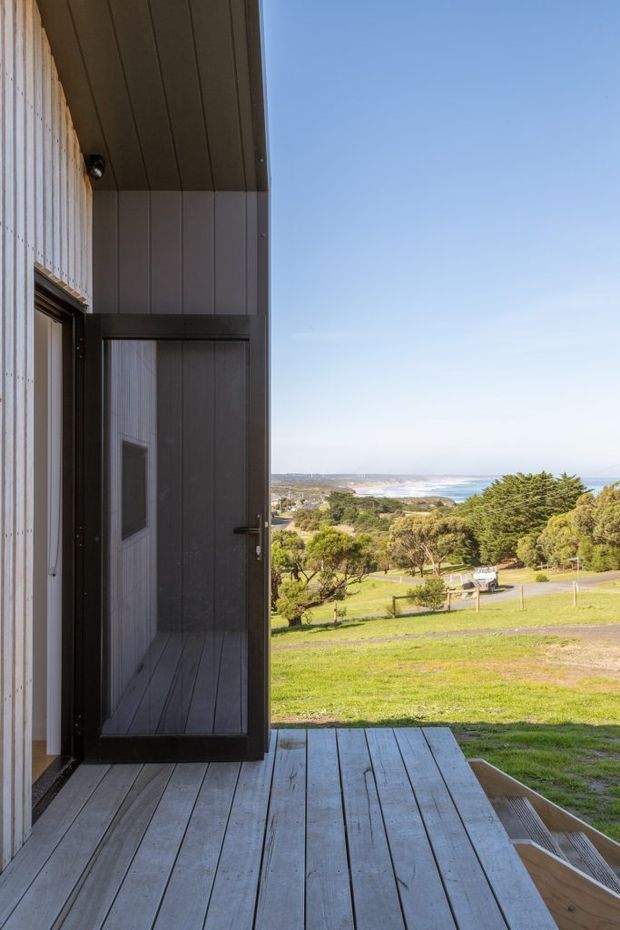 A vertical exterior view from a raised timber deck, looking past an open dark-framed doorway toward a landscaped coastal property and distant ocean shoreline. The building has dark vertical cladding, a timber-lined soffit, and weathered grey decking, with expansive blue sky and a bright coastal outlook.