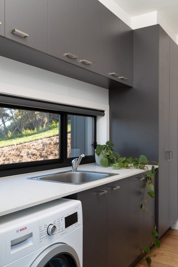 Compact, contemporary laundry with dark grey floor-to-ceiling cabinetry, a white benchtop, stainless-steel sink and tap, front-loading washing machine, black-framed window, and timber flooring. A trailing indoor plant sits beside the sink, with a leafy outdoor view through the window.