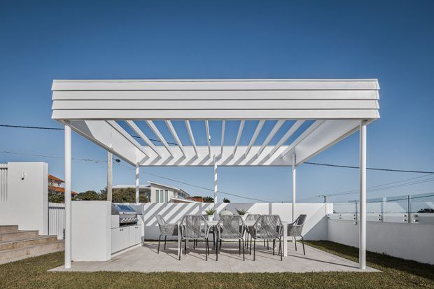 A bright contemporary outdoor dining area beneath a white slatted pergola. The covered patio has a dining table with woven chairs, a built-in outdoor barbecue or kitchen unit, white rendered boundary walls, and a low-maintenance lawn. Strong linear shadows from the pergola create a crisp geometric effect against the pale paving and blue sky.