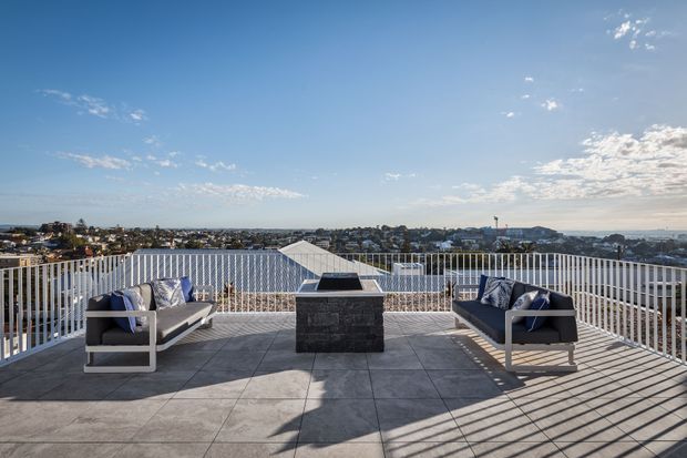 A spacious rooftop terrace or elevated outdoor deck with a tiled floor, white vertical balustrade, two contemporary outdoor lounge chairs, and a central dark stone-clad fire pit or feature table. The terrace overlooks a broad suburban coastal or urban landscape under a bright blue sky.