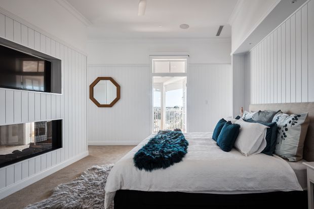 Bright, white coastal-style bedroom with vertical wall panelling, a large upholstered bed, layered white and deep teal bedding, a shag rug, and a built-in double-sided fireplace and television wall. An octagonal timber-framed mirror and balcony doorway add architectural interest, while daylight enters through the open glazed door.