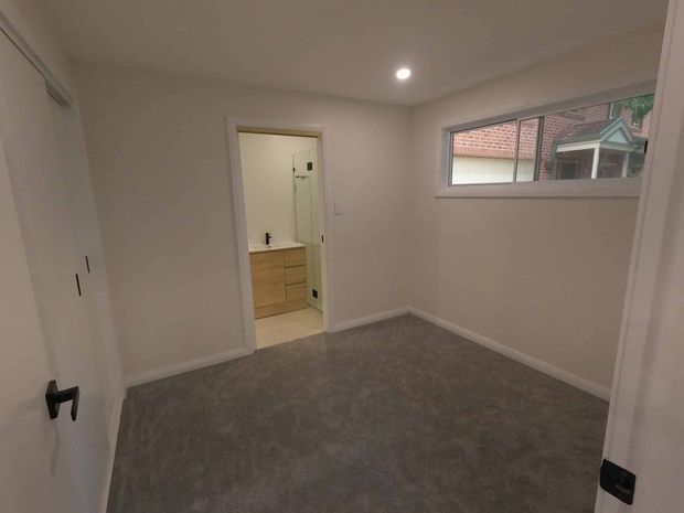 A small, empty carpeted bedroom with off-white walls, built-in white wardrobes, recessed ceiling lighting, and a wide horizontal window. An open doorway leads directly to a bathroom or ensuite area with a timber-toned vanity and mirror.