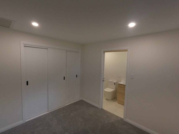 A small, newly finished bedroom with grey carpet, white walls and recessed ceiling lights. Built-in white sliding-door wardrobes occupy one wall, and an open doorway reveals an adjoining bathroom with a toilet and light timber vanity.