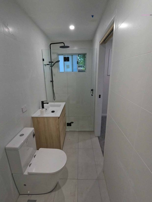Compact contemporary bathroom with white tiled walls and floor, a wall-to-wall glass shower enclosure with matte black shower fittings, a light timber vanity with white basin, and a white toilet. A small window provides natural light at the rear.