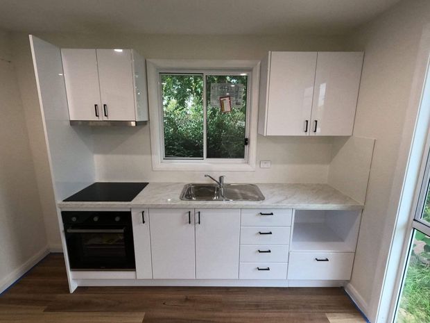 Compact, bright contemporary kitchen with white gloss cabinetry, a pale stone-look benchtop, stainless-steel sink and tap, black cooktop, built-in oven, and dark timber-look flooring. A central window provides natural light and views of greenery.