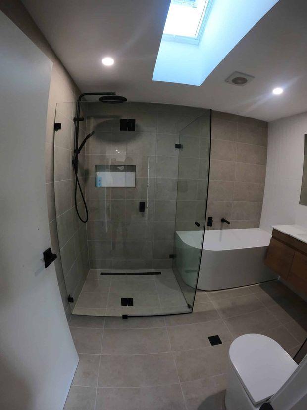 Modern bathroom featuring a large frameless glass walk-in shower with black hardware, a freestanding white bathtub, wall-to-wall grey-beige tiled surfaces, a timber vanity, skylight, recessed lighting, and a partially visible toilet.