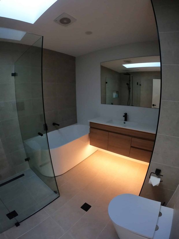 Contemporary bathroom with a frameless glass shower enclosure, freestanding white bathtub, wall-to-wall mirror, floating timber vanity with integrated basin, dark tapware, tiled walls and floor, and warm under-vanity lighting. A toilet is visible in the foreground.