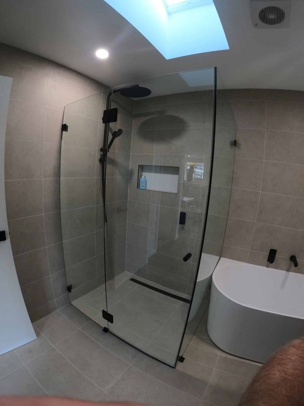 Modern bathroom featuring a large frameless glass walk-in shower with black fixtures, a freestanding white oval bathtub, large beige-grey wall and floor tiles, recessed ceiling lighting, and a skylight.