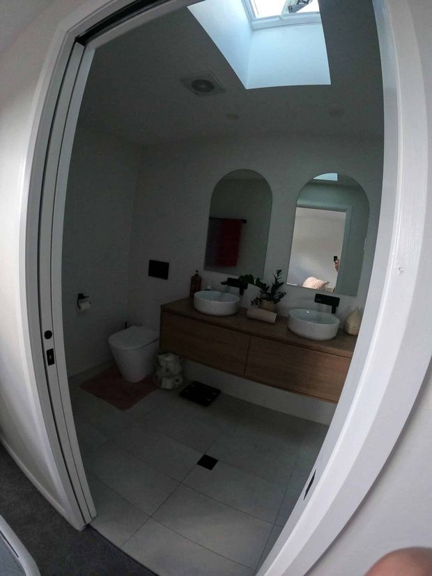 A contemporary bathroom viewed through an open doorway, featuring a floating timber vanity with two white vessel basins, arched wall niches or mirrors, a toilet, large light-grey floor tiles, and a skylight providing natural illumination. The room has white walls and a clean, minimal fit-out.