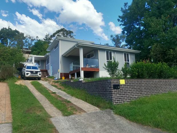 Exterior street-level view of a raised single-storey house with light grey horizontal weatherboard cladding, a low-pitched roof, covered front porch with timber decking and glass balustrade, and a dark brick retaining wall along the front boundary. A concrete driveway and landscaped garden surround the home.