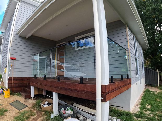 Exterior view of a raised, single-storey home addition or porch with grey horizontal weatherboard cladding, white trim and posts, a low-pitched roof, and a timber-framed deck enclosed by clear glass balustrading. The elevated deck and open subfloor are prominent, with a side garden and fencing visible.