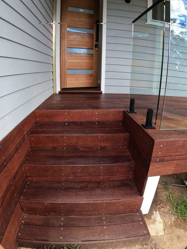 Exterior view of a raised entry porch with a short flight of dark timber stairs and matching timber decking. The landing leads to a timber front door with horizontal frosted-glass inserts. Light horizontal weatherboard cladding surrounds the entry, with a clear glass balustrade and dark metal fittings on the right. The image focuses on the outdoor access and facade details.