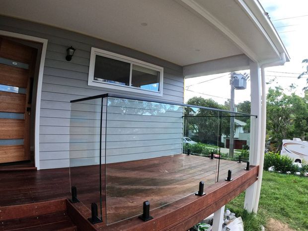 A covered raised front deck and entry porch with dark timber decking, clear glass balustrading in black brackets, white posts and trim, and a grey horizontal weatherboard exterior. A timber front door and small window are visible, with suburban surroundings beyond.