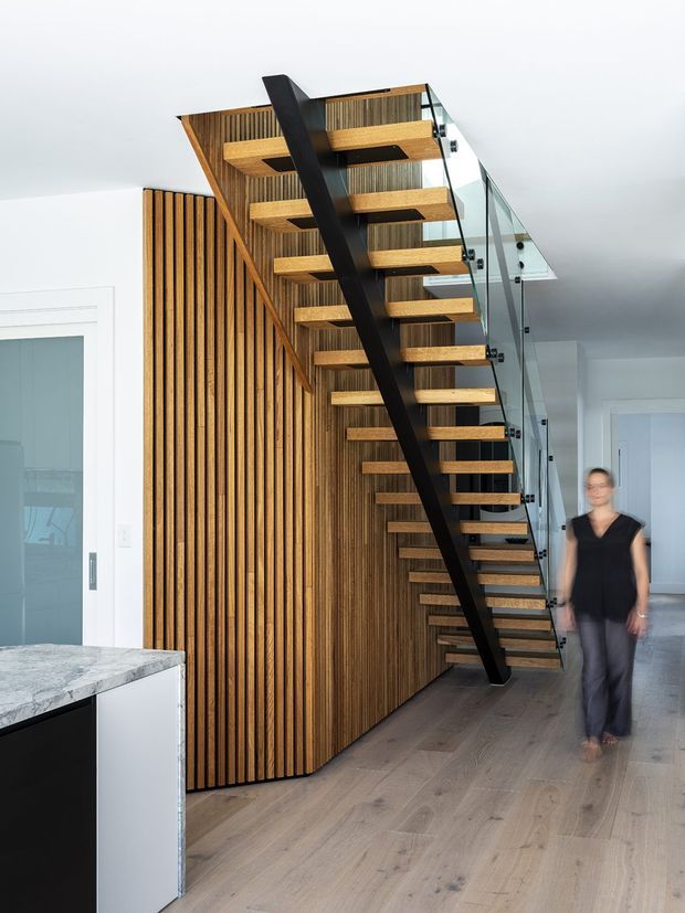 Interior view dominated by a contemporary open-riser staircase with thick timber treads, a black steel central stringer, and clear glass balustrades. The staircase rises beside a full-height vertical timber slat feature wall and appears to lead to an upper landing or mezzanine. Pale timber flooring, white walls, and a partial stone-topped kitchen island are visible in the foreground.