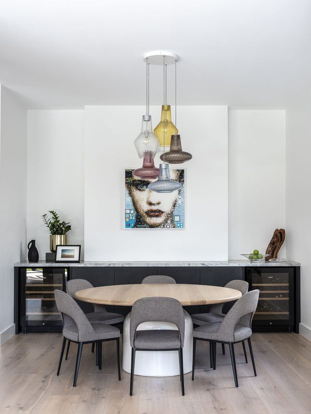 A refined contemporary dining room with a large oval timber dining table surrounded by upholstered grey dining chairs. A long dark built-in sideboard with integrated glass-front wine storage runs along the rear wall beneath a marble-look benchtop. A colourful mosaic portrait provides a focal point, while a cluster of sculptural pendant lights hangs above the table. The room has white walls, pale timber flooring, and a clean, balanced composition.