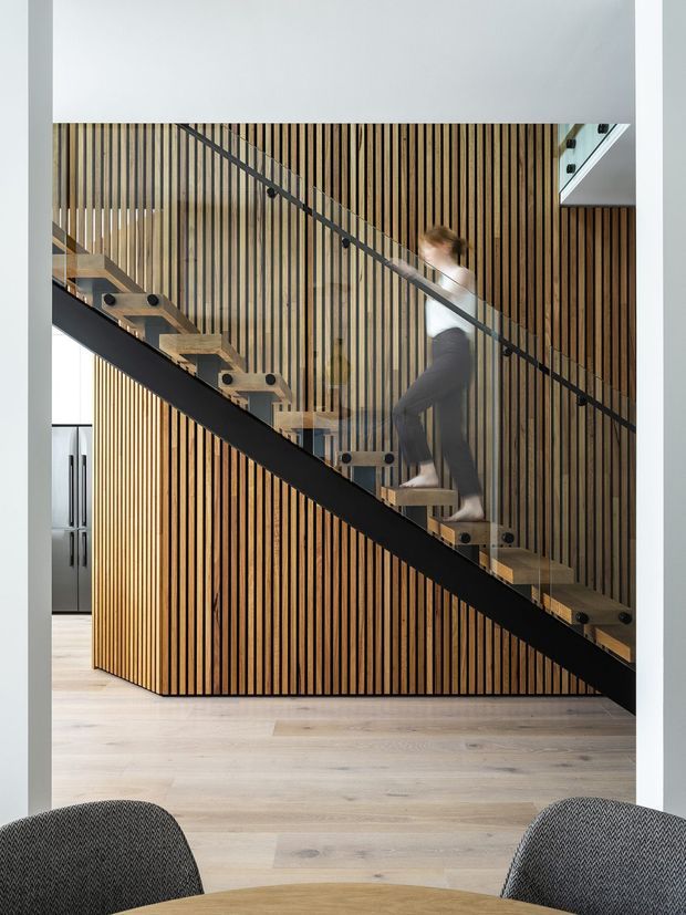 Interior view focused on a contemporary open-riser staircase with clear glass balustrades, black structural stringers and handrails, and timber treads. The stair is set against a striking wall of closely spaced vertical timber battens, with pale timber flooring and partial dining chairs in the foreground. A glimpse of a kitchen is visible to the left. The composition is clean, warm, and architectural, with a person ascending the stairs.