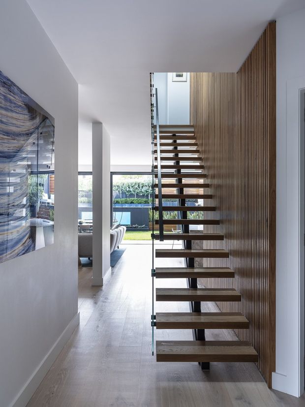 A bright contemporary interior hallway centered on a floating open-tread staircase. The staircase has thick dark timber treads supported by slim dark metal elements and clear glass side panels, set against a full-height vertical timber slat feature wall. Pale timber flooring, white walls and ceiling, and strong glazing create an airy connection to the living area and landscaped exterior beyond. A large abstract artwork is visible on the left wall.