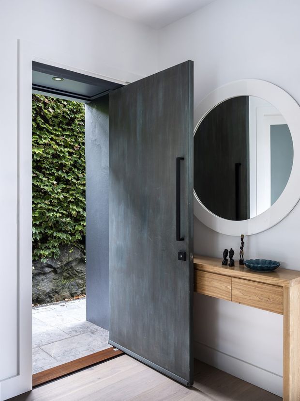 A refined residential entryway viewed from inside, featuring a tall dark timber or metal-look pivot door opening to a leafy exterior, a round wall mirror, and a slim light-wood console with drawers and decorative objects. The space has white walls, pale timber flooring, recessed lighting, and a strong contrast between the dark door and light finishes.