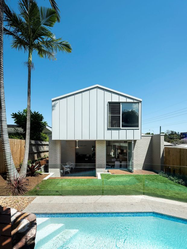 A compact two-storey contemporary house viewed from the swimming pool. The upper level has a simple gabled form clad in pale vertical metal panels with a large framed window, while the lower level is partially open with light brick columns, extensive glazing and a small terrace. A neatly fenced yard with artificial lawn, palms and landscaping sits between the house and the bright blue pool.