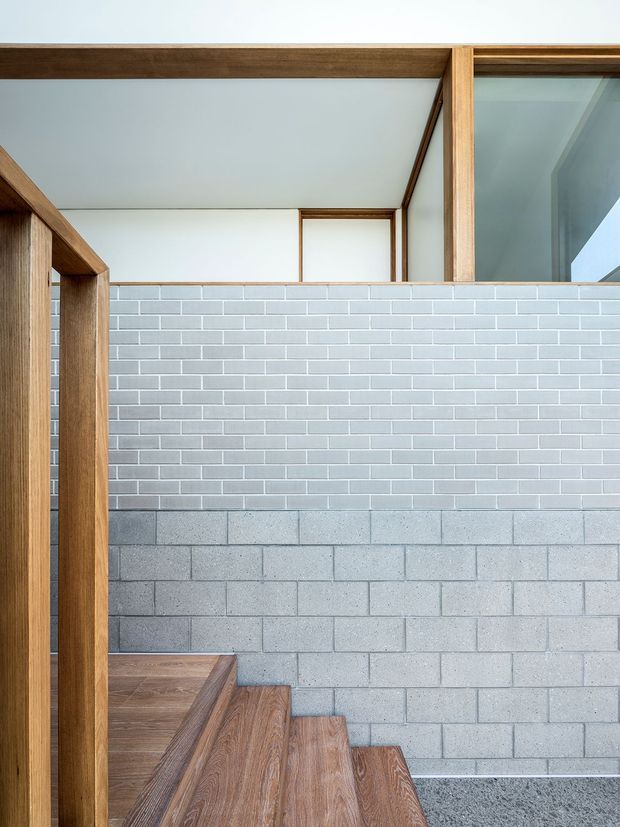 A contemporary interior stairway with warm timber treads and framing, rising beside a two-tone masonry wall. The lower wall is formed from exposed grey concrete block, while the upper section has small pale grey glazed brick tiles. A large internal window or glazed opening and white ceiling add light and openness to the minimalist composition.