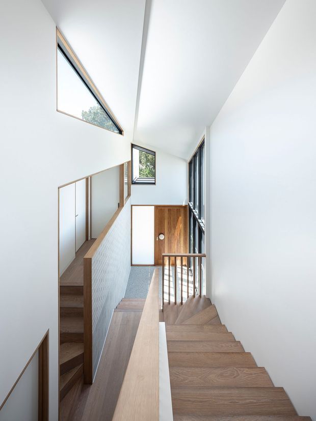 Bright, narrow multi-level stairway with white walls and ceiling, warm timber stair treads, timber handrails and balustrades, a timber entry door, and tall windows providing natural light. The stairwell has a clean contemporary design with strong vertical and diagonal geometry.