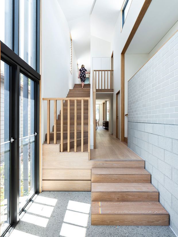 Bright contemporary interior view of a multi-level stairway and landing. Wide timber stairs with matching timber balustrades rise through a tall open stairwell, bordered by large black-framed glazed openings on the left and a white painted masonry feature wall on the right. Pale timber flooring, white walls, exposed timber trim, and strong daylight create an airy, clean vertical circulation space.