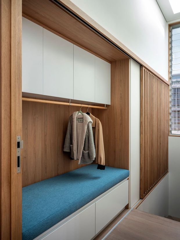 Built-in entryway storage with a timber-lined coat nook, white overhead cupboards, hanging rail, bright blue upholstered bench seat, and integrated drawers below. A vertical timber slat screen and louvred window bring filtered daylight into the compact, carefully detailed space.