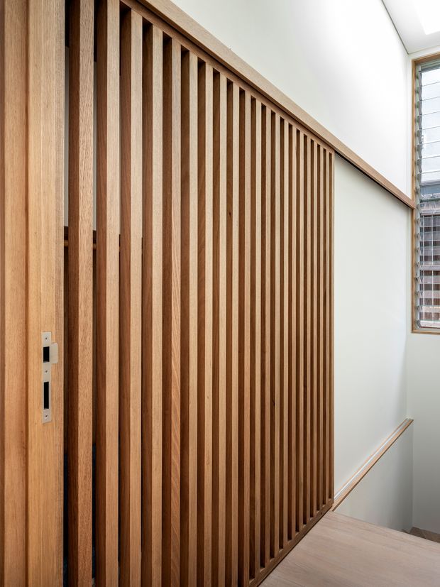 Interior view of a stair landing featuring a full-height vertical timber slat screen and matching timber handrail. The warm wood partition conceals the stair opening, while white walls, a pale timber floor, and a louvred window provide a clean, contemporary architectural backdrop.