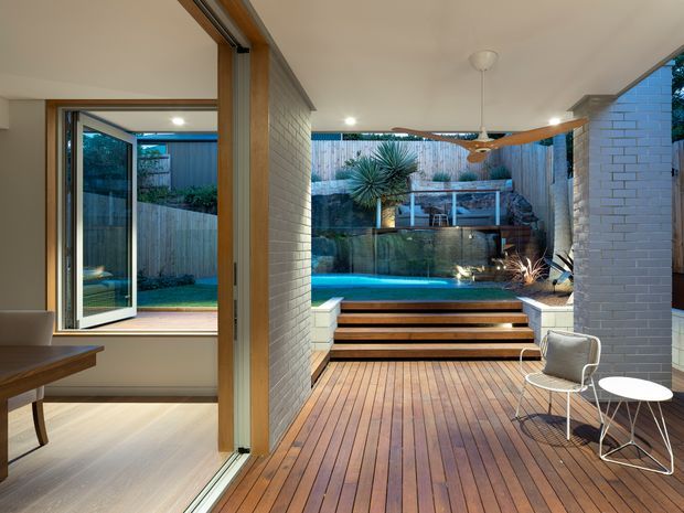 A contemporary indoor-outdoor living area opening onto a timber deck and illuminated swimming pool. The space features white-painted brick walls, large timber-framed bi-fold glazing, warm timber decking and steps, minimalist outdoor furniture, integrated garden planting, and a sculptural ceiling fan. A small interior room is visible through the open doors on the left.