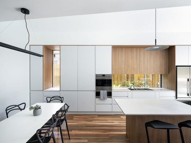 A bright, open-plan contemporary kitchen and dining area with a long white dining table and black wire-frame chairs in the foreground. The kitchen features full-height white cabinetry, warm timber cabinetry and timber flooring, integrated appliances, a white island benchtop with timber base, and a horizontal window above the sink. Minimal black pendant and track lighting add contrast.