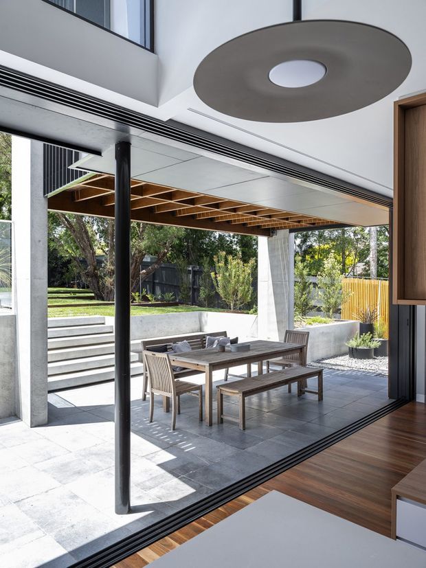 A bright contemporary indoor-outdoor dining area viewed from inside through fully opened glazed doors. The space features a timber dining table with bench seating and chairs on a large grey tiled patio, framed by concrete columns and a black slender structural post. Above, a timber pergola with exposed beams provides shade, while steps, raised garden beds, fencing and mature planting form the outdoor setting. Inside, dark timber flooring, white walls, a built-in timber cabinet and a large circular ceiling light are visible.