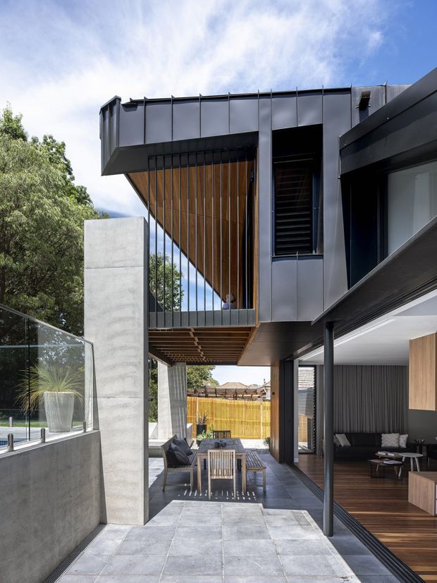 A contemporary multi-level residential exterior and covered outdoor living area. The facade combines dark metal cladding, exposed concrete walls, timber soffits and vertical timber screening, with extensive glazing and slim black framing. A paved courtyard or terrace contains outdoor dining furniture, while an open living room is visible to the right. A glass pool barrier and landscaped greenery appear at the edge of the frame.